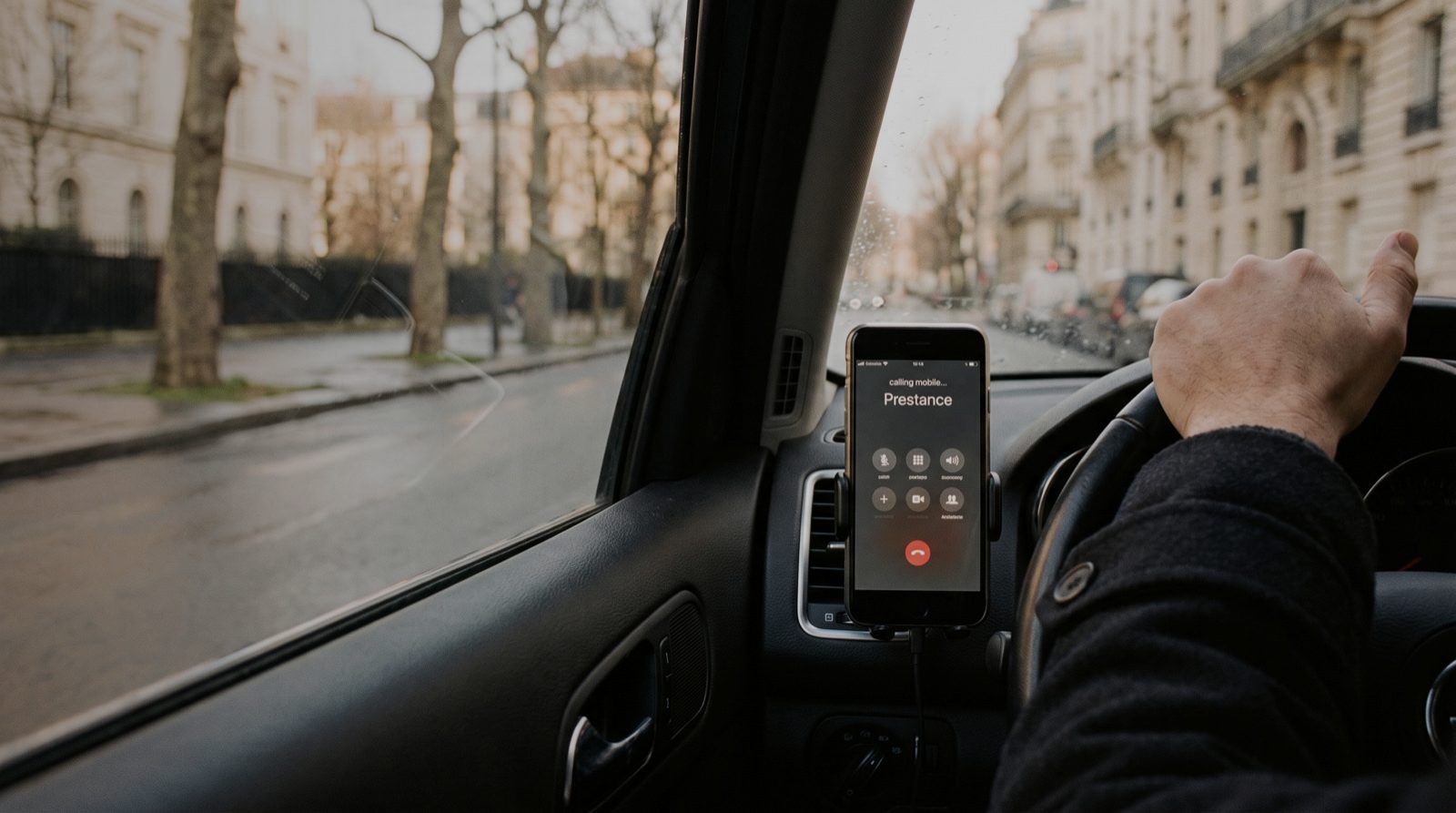 A phone in a dashboard cradle, seen from the driver seat: the screen shows an outgoing call in progress to Prestance, with the speaker, mute and end-call controls below it. A forearm rests on the steering wheel at the edge of the frame; through the windscreen, a blurred street of pale stone buildings and bare trees on an overcast afternoon.