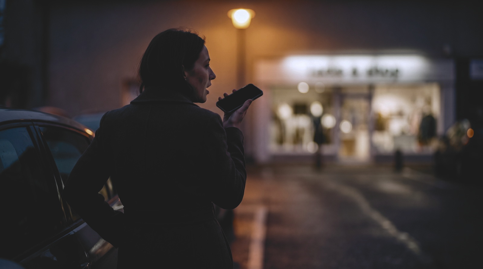 A woman in a dark tailored coat stands beside a car in a boutique car park at dusk, seen from behind and in profile. She holds a phone near her mouth and is speaking; the phone screen is dark and blank. A street lamp casts warm orange light on the wet tarmac, and a lit shop front glows out of focus behind her.